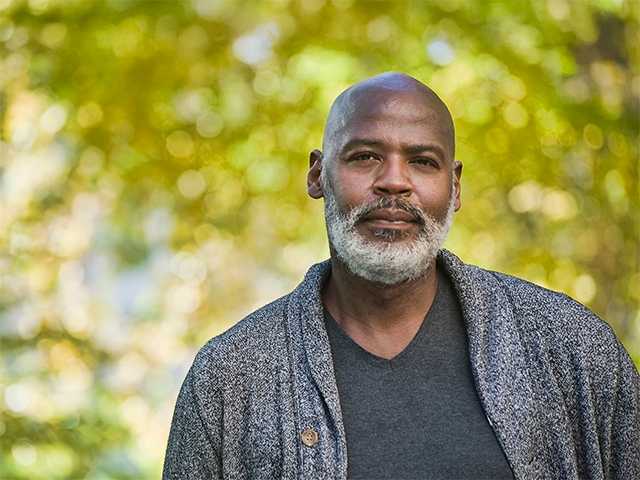 Black man with a grey beard and wearing a black sweater stands outside in the sunshine