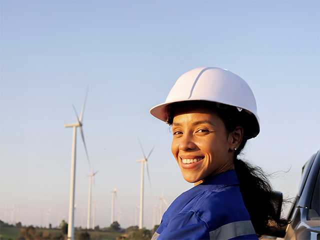 Smiling woman with a ponytail and in a blue jumpsuit stands in front of wind turbines outdoors