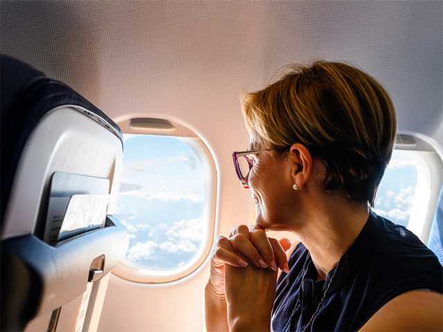 A woman peers out the window of an airplane, watching the clouds and scenery as she travels through the sky.