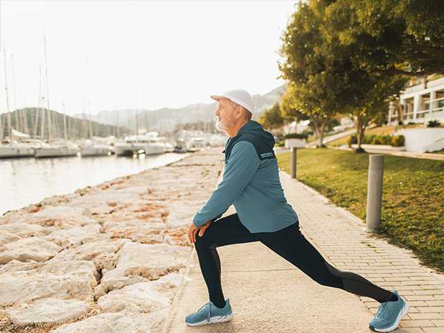 Older man wearing black jogging pants and a blue jogging jacket does a lunge stretch while outside