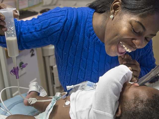 Young Black woman in a blue shirt leans over and smiles at her baby in the neonatal intensive care unit