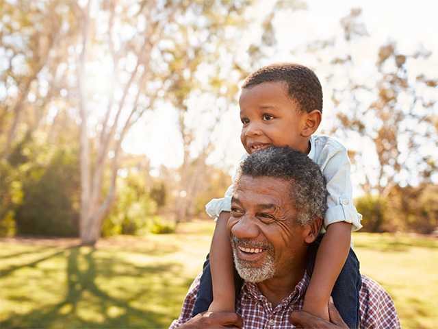 Elderly Black man carries his grandson on his shoulders outdoors in the sunshine