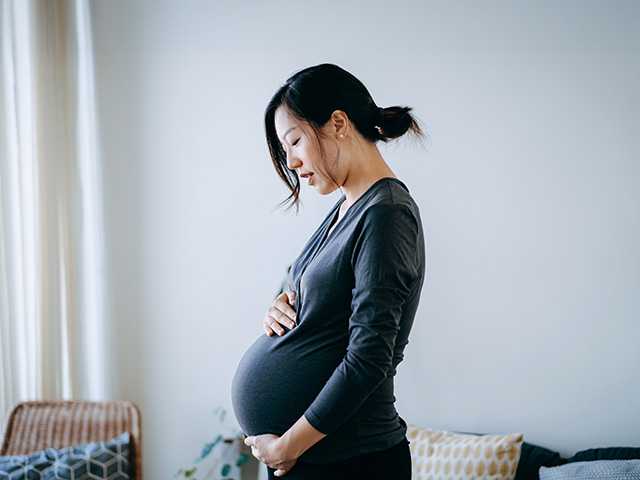 Pregnant Asian woman stands in a sunlit room with her hands on her belly