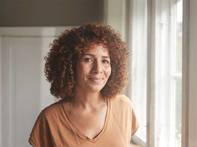 Smiling Black woman with curly brown hair stands in a sunny room