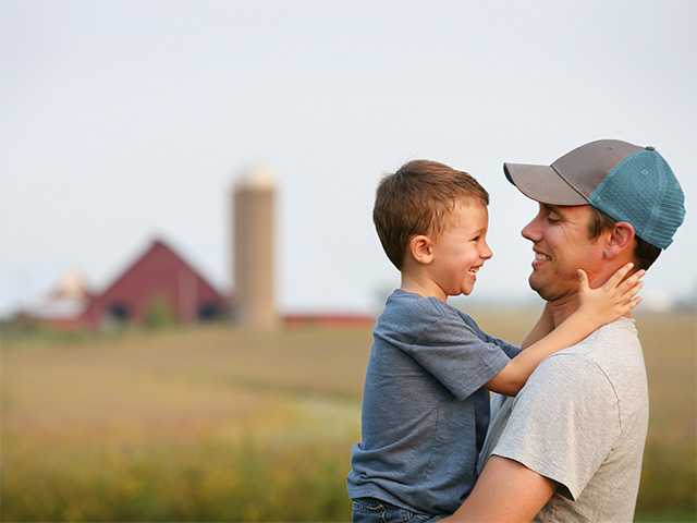 Smiling young white man lifts up his young son while they stand in a field with a barn and silo in the background