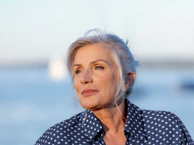 Older white woman with grey hair stands in front of a lake in the evening sun