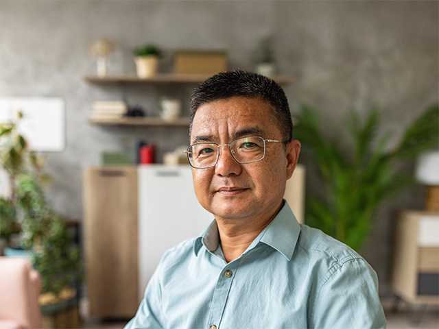 Middle aged Asian man in a blue shirt and wearing glasses sits in a room with plants behind him