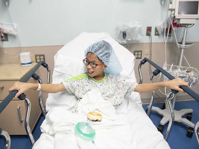 Young boy wearing glasses and surgical gown sits in hospital bed