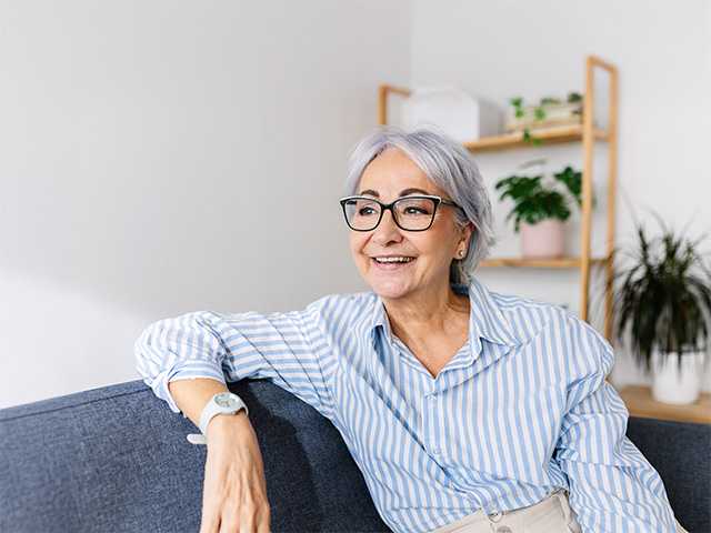 White woman with grey hair and glasses sitting on a sofa and resting her arm on the back of it