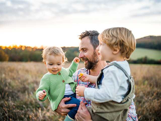 Middle aged man holds two toddlers as he stands in a field in the evening