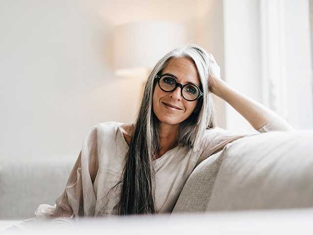 Smiling older white woman wearing glasses rests her elbow on a couch