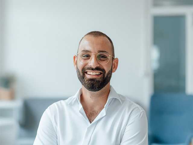 Smiling young white man with glasses and a beard inside a white room