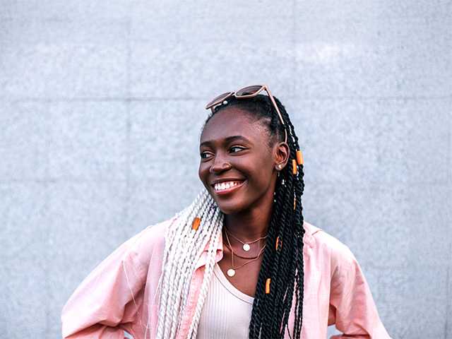 Smiling young Black woman with braided hair stands outdoors in front of a grey wall